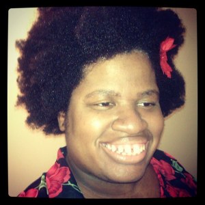 An Instagram image that is square shaped with a black border. Image of a Black woman with curly natural hair with a small red bow on the left side of her hair. She is wearing a black long-sleeved shirt with a print of red flowers. She is smiling at the camera and looking slighting off camera.