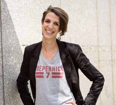 Photo of a white woman, smiling and standing with her hands on her hips and head slightly tilted to the left, in front of a gray granite wall. She has short brown hair in an asymmetrical haircut, longer on her left side and shorter on her right side. She is wearing dangling silver earrings and a gray t-shirt that reads "Kaepernick 7" in red, underneath a black blazer.