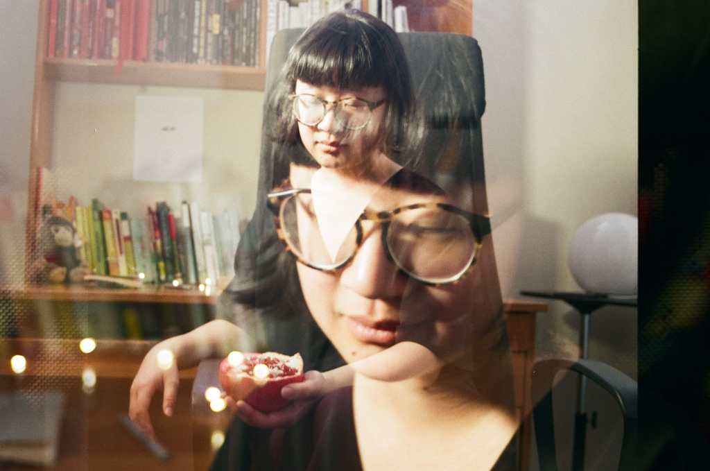 A double exposure photograph, wherein two images are faded onto one another in a single image, of Jane sitting at her desk on a black office chair. Jane is a light skin East Asian person, with her eyes closed, wearing brown and black glasses and short black hair that slightly flares at the end with rounded bangs like a bowl. In one exposure Jane’s face is larger and closer to the camera; there are lights directly behind her. In the other, she is sitting on a black office chair and holding a sliced open half of a pomegranate. Behind her is a round, moon-like light. The desk behind her is a medium orange-brown and contains a head shelf, with books ordered in red, black, white, and the rest of the rainbow ordered on the top of the table. There’s a small brown monkey plushie near the left end of the desk. There’s a black and white ink illustration above the orange to blue books on top of the desk where there’s an open space.