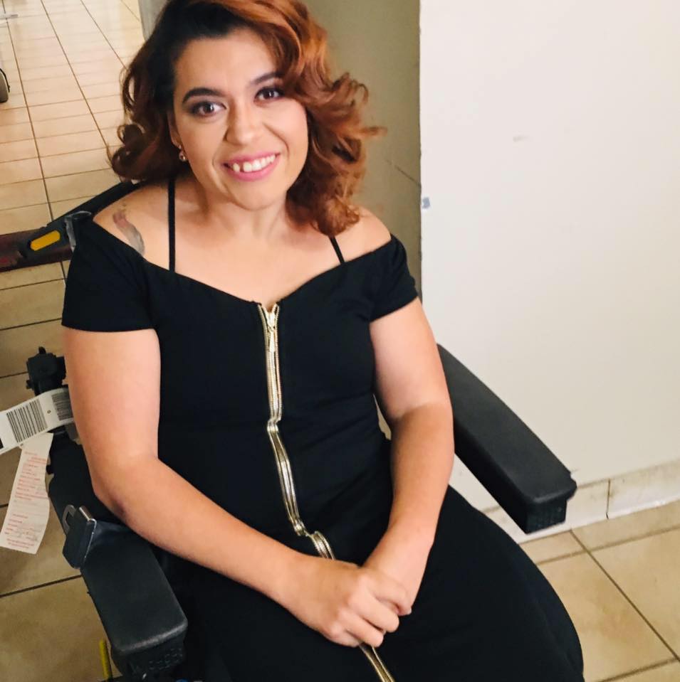 Una foto de Ashley, mujer filipina/blanca de piel aceitunada con el pelo rizado castaño rojizo hasta los hombros, sonriendo con la cabeza ligeramente hacia un lado. Está sentada en una silla de ruedas eléctrica con un vestido de cóctel negro con una cremallera dorada que le llega hasta la mitad y las manos cruzadas en el regazo