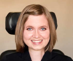 Headshot of a young white woman with blue eyes with chin-length golden brown hair. She is in a wheelchair and a headrest is behind her.