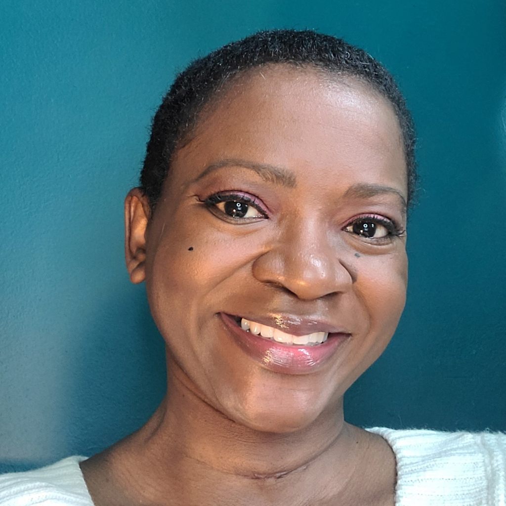 Photo of Carolyn Hinds, a Black female with dark brown complexion, close cropped black hair, wearing a wide smile, and white sweater, in front of a teal colored wall.