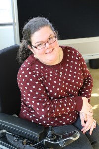 Young white woman in a wheelchair. Her curly brown hair is tied back. She is wearing eyeglasses and a maroon sweater with white polka dots. Her wheelchair is black and in the background is a chalkboard.