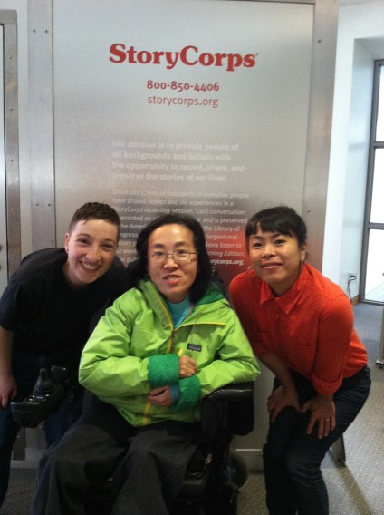 Image description: he photo is of three people in front of a StoryCorps booth. The person on the left is Yosmay del Mazo. He is Latinx and Transgender, is wearing a black shirt, has short dark hair and is smiling at the camera. The person in the middle is Alice Wong. She is an Asian American woman, and she’s wearing a green rain coat and glasses. She’s smiling at the camera and is in a power chair. The person on the right is Geraldine Ah-Sue. She is an Asian American woman, and she’s wearing a red button-up shirt and black pants. She’s smiling at the camera.