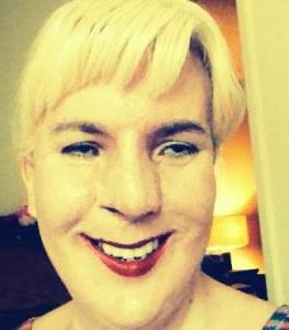 Image of Jennifer Justice, a white woman with short blonde hair. She is wearing bright red lipstick and is smiling at the camera. Her image has a filter that gives a yellow tint.