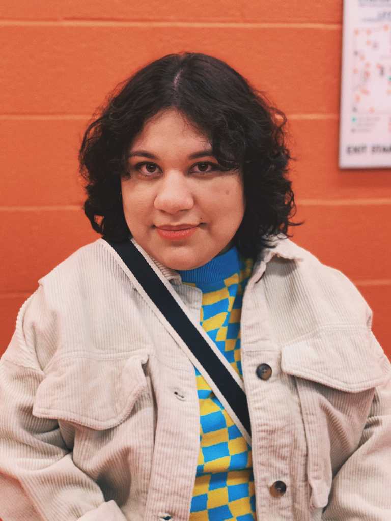 Photo of Jocelyn Mondragon, a Mexican woman with curly short black hair, smiling straight at the camera. In front of an orange painted brick wall, Jocelyn is wearing a beige corduroy jacket with a blue and yellow checkered sweater underneath. A black and white striped strap is across her shoulder.