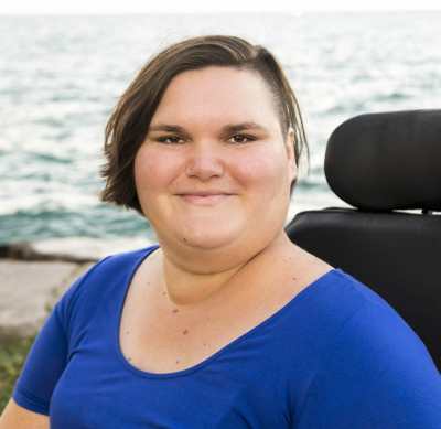 A headshot of Kennedy, a white, fat, femme, smiling softly at the camera. She has chin length brown hair with one side shaved and is wearing a blue shirt. Her electric wheelchair and Lake Michigan are behind her.