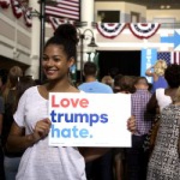 Photo of a young woman of color with a white t-shirt on holding a sign that says, "Love trumps hate" at a political rally. Behind her are crowds and a stage.