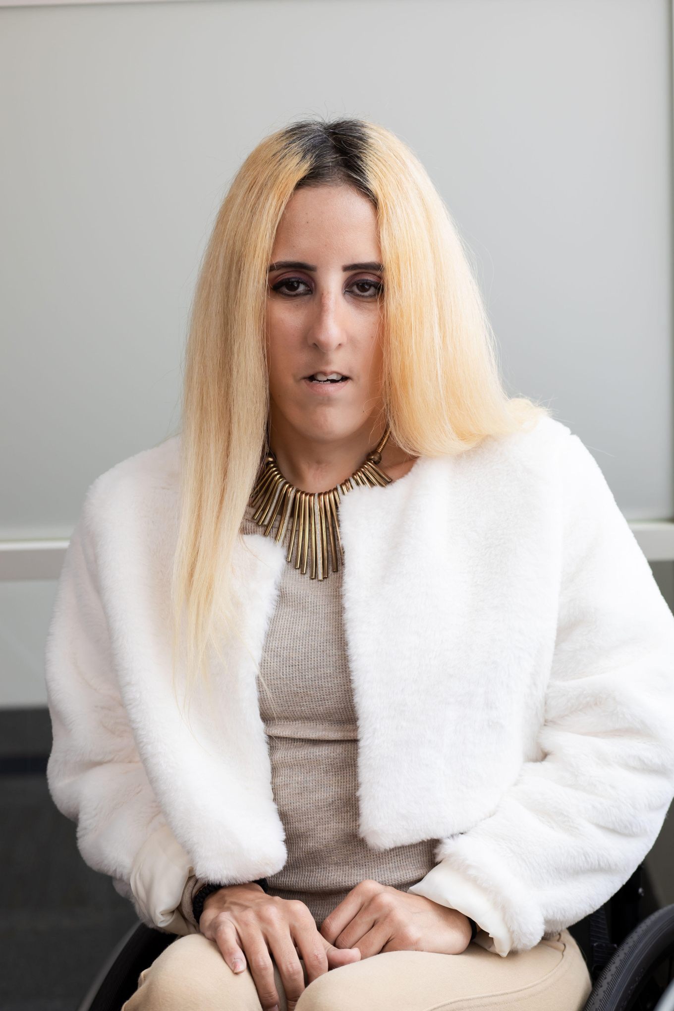 A headshot of a woman with brown color eyes and olive-tan skin complexion with long, straight blonde hair, wearing a baige top with a white jacket and a gold necklace.