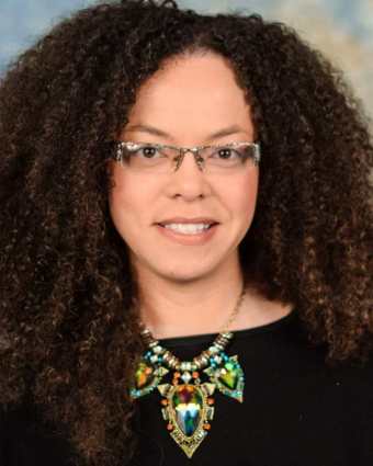 Headshot of a light complected African American woman, late thirties, with curly brown hair and glasses. She is wearing a black top and a large statement necklace
