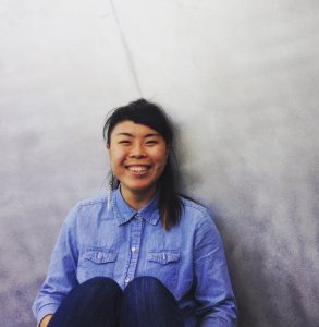 An Asian American woman of color with long black hair tied up in a ponytail sitting in front a light gray metal sculpture. She is wearing a denim blue button up shirt and jeans, and she is smiling.