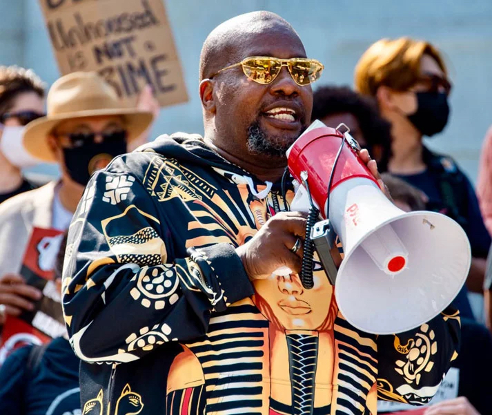 Theo Henderson, a Black man with a shaved head, goatee, gold-tinted sunglasses, and wearing a hoodie with an image of Tutankhamun on it, speaks into a red and white megaphone at a protest. Behind Theo is a crowd of people wearing face masks, with one holding up a sign that says "Being unhoused is not a crime." Credit: WeTheUnhoused.com