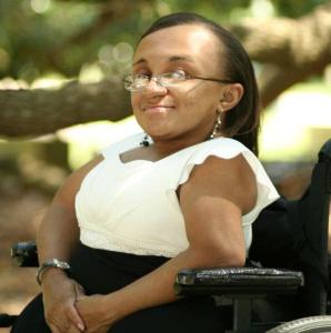 Photo of a young Black woman with her hair parted and hanging down straight. She is smiling and facing the camera sideways in her wheelchair. She is outside under the tree branches while wearing a white and black dress.
